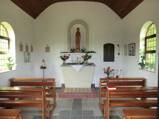 Kleiner Kapellenraum mit weißem Altar und Madonnenfigur, umgeben von Holzbänken und schlichten Dekorationen.
