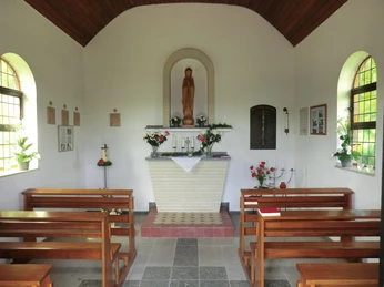 Heimkehrerkapelle Innenraum Kleiner Kapellenraum mit weißem Altar und Madonnenfigur, umgeben von Holzbänken und schlichten Dekorationen.