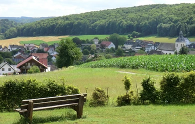 Teutoburger-Wald - Brakel Blick von der Heimkehrerkapelle auf Istrup Holzbank mit Blick auf ein Dorf im Tal, umgeben von grünen Feldern und Wald im Hintergrund.
