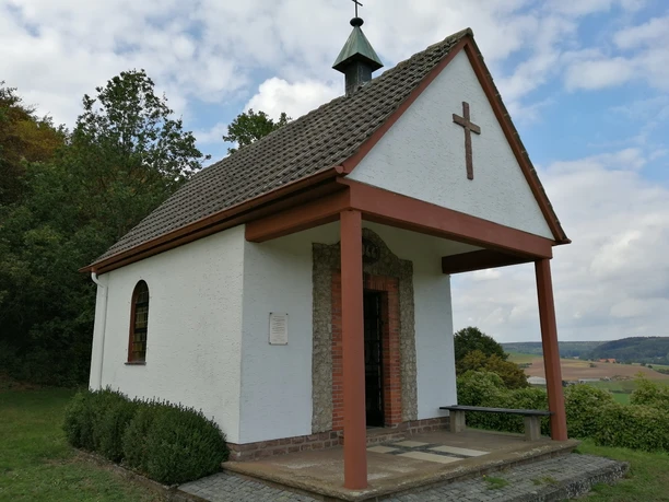 Weiße Kapelle mit schiefergedecktem Satteldach und hölzernem Vordach in ländlicher Landschaft.