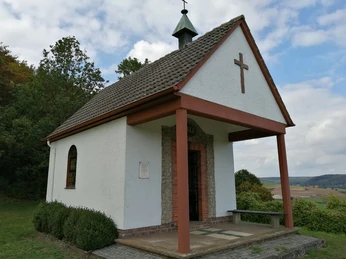 Heimkehrerkapelle - Außenansicht Weiße Kapelle mit schiefergedecktem Satteldach und hölzernem Vordach in ländlicher Landschaft.