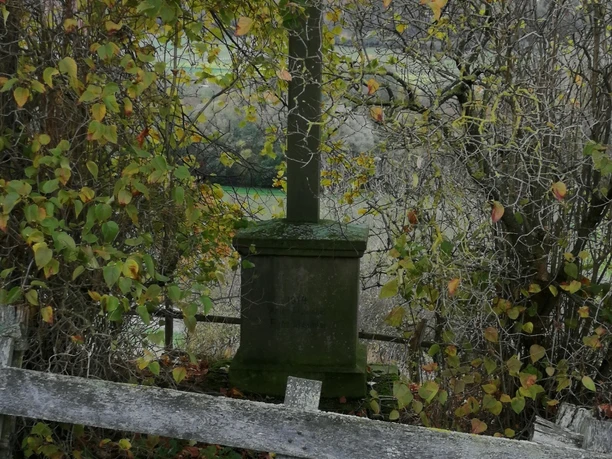 Krawinkels Kreuz Ein steinernes Kreuz steht inmitten umgebenden Herbstlaubes, flankiert von einem Holzzaun.