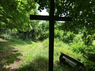 Schattiges Holzkreuz unter Bäumen an einem Waldweg, daneben eine Holzbank für eine stille Pause.