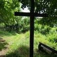 Schattiges Holzkreuz unter Bäumen an einem Waldweg, daneben eine Holzbank für eine stille Pause.