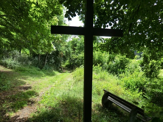 Winklers Kreuz Schattiges Holzkreuz unter Bäumen an einem Waldweg, daneben eine Holzbank für eine stille Pause.