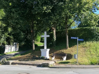 Weißes Steinkreuz auf kleinem Hügel, umgeben von Bäumen, Bank und Straßenschildern in grüner Umgebung.
