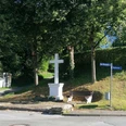 Bartholomäus Kreuz Weißes Steinkreuz auf kleinem Hügel, umgeben von Bäumen, Bank und Straßenschildern in grüner Umgebung.