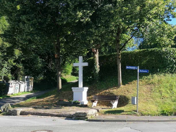 Bartholomäus Kreuz Weißes Steinkreuz auf kleinem Hügel, umgeben von Bäumen, Bank und Straßenschildern in grüner Umgebung.