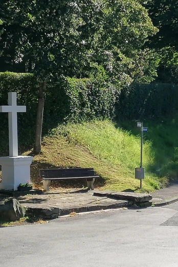 Steinernes Kreuz mit Bank an einer von Bäumen gesäumten Straße, umgeben von grüner Natur.
