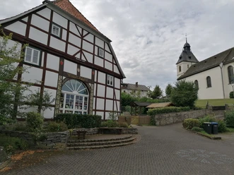 Fachwerkhaus und Kirche St. Bartholomäus mit Turm und Garten, umgeben von Bäumen und gepflastertem Platz.