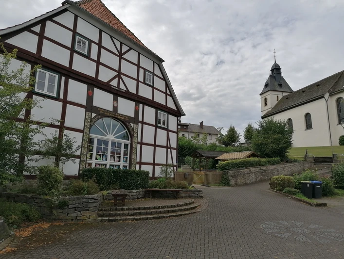 Pfarrheim mit Pfarrkirche St. Bartholomäus Fachwerkhaus und Kirche St. Bartholomäus mit Turm und Garten, umgeben von Bäumen und gepflastertem Platz.