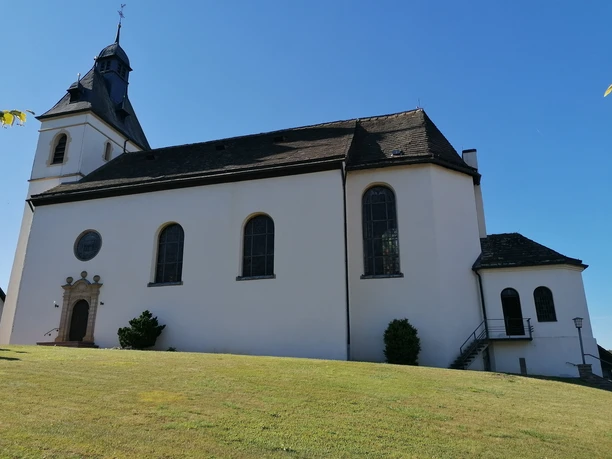 Weiße Kirche mit dunklem Turmdach und großen Fenstern auf einer leicht abschüssigen grünen Wiese.
