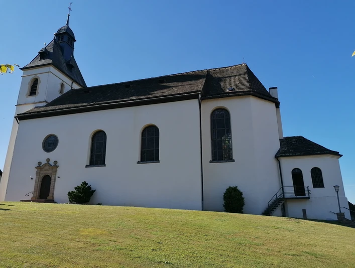 Weiße Kirche mit dunklem Turmdach und großen Fenstern auf einer leicht abschüssigen grünen Wiese.