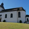 Pfarrkirche St. Bartholomäus Weiße Kirche mit dunklem Turmdach und großen Fenstern auf einer leicht abschüssigen grünen Wiese.