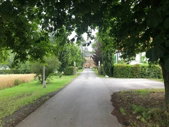 Ellerburg Aue Radweg - 2. Tour Blick auf einen von Bäumen gesäumten Weg, der zu einem Backsteingebäude inmitten grüner Vegetation führt.