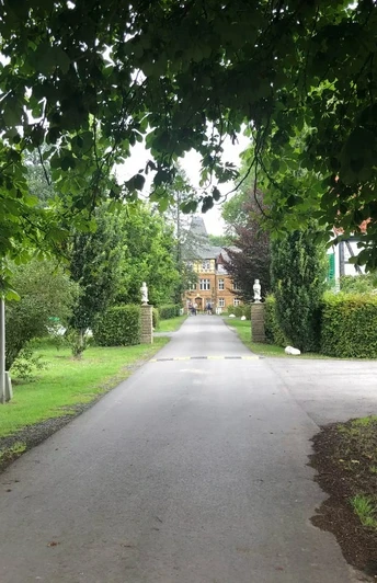 Ellerburg Aue Radweg - 2. Tour Blick auf einen von Bäumen gesäumten Weg, der zu einem Backsteingebäude inmitten grüner Vegetation führt.