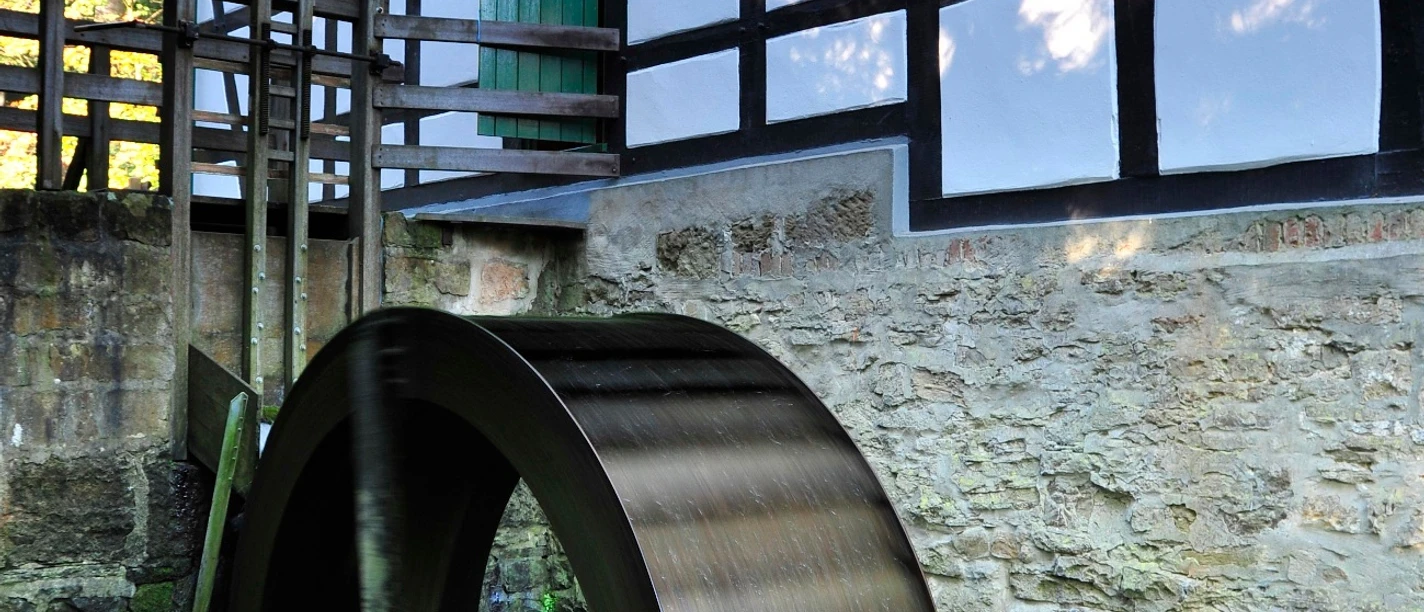 Historische Wassermühle Hudenbeck mit beeindruckendem Fachwerk und rotierendem Wasserrad im Grünen.