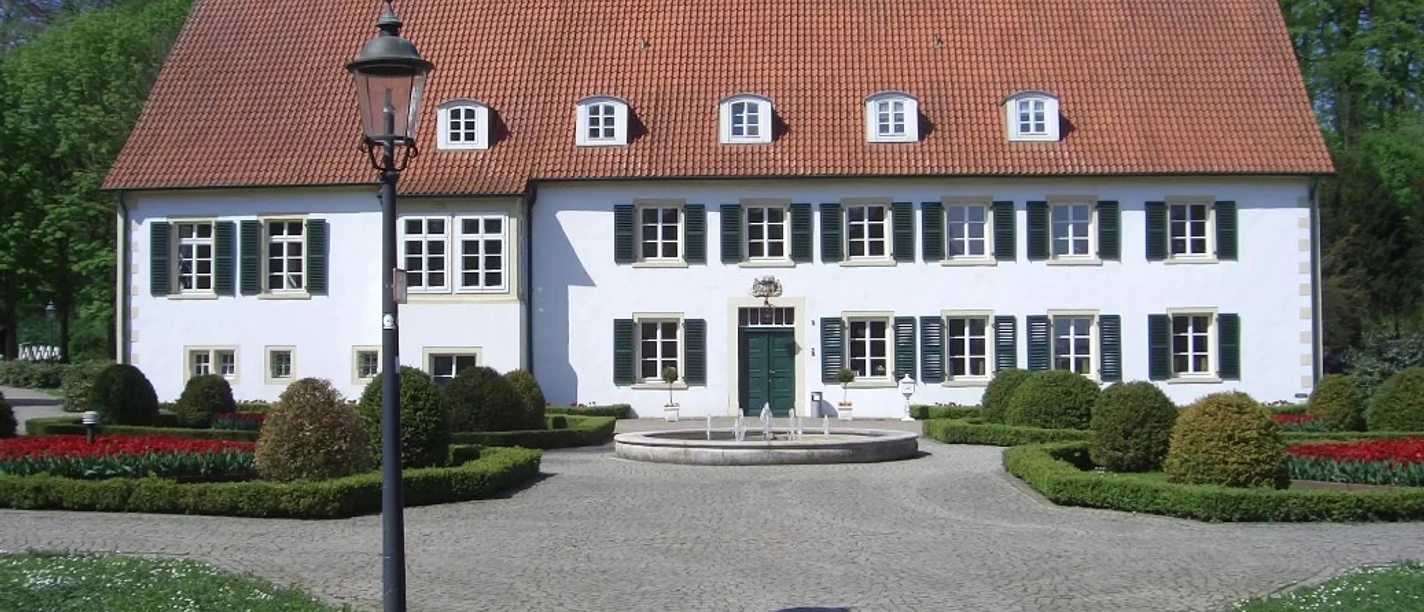 Historisches Gebäude mit rotem Ziegeldach, umgeben von gepflegten Gärten und Kopfsteinpflasterweg.