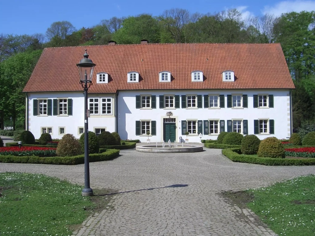 Haus des Gastes Bad Holzhausen Historisches Gebäude mit rotem Ziegeldach, umgeben von gepflegten Gärten und Kopfsteinpflasterweg.