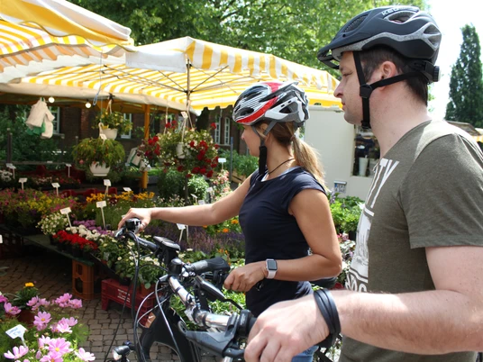 Achimer Wochenmarkt Zwei Radfahrer mit Helmen betrachten einen Marktstand voller bunter Blumen auf einem Wochenmarkt.