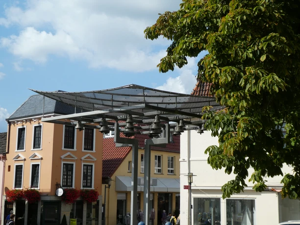 Achimer Glockenspiel Eine moderne Glockenspielinstallation vor historischen Gebäuden mit Blätterdach im Vordergrund.