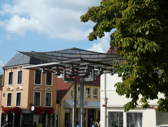 Achimer Glockenspiel Eine moderne Glockenspielinstallation vor historischen Gebäuden mit Blätterdach im Vordergrund.