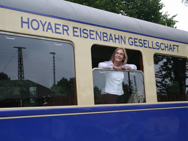 Frau in einem nostalgischen, blau-beigen Zug der Hoyaer Eisenbahn Gesellschaft vor grünen Bäumen.