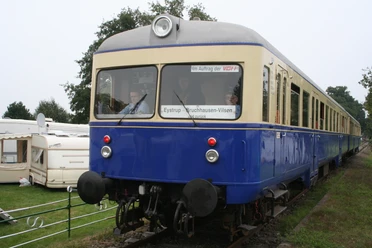 Ein blauer nostalgischer Zug auf Gleisen, umgeben von grüner Landschaft und einem Wohnwagen.
