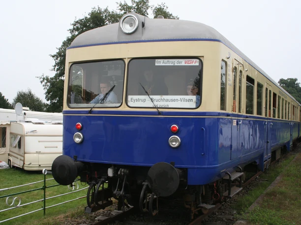 Kaffkieker Ein blauer nostalgischer Zug auf Gleisen, umgeben von grüner Landschaft und einem Wohnwagen.