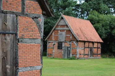 Zwei historische Fachwerkgebäude mit Backsteinwänden stehen auf einer grünen Wiese vor Bäumen.