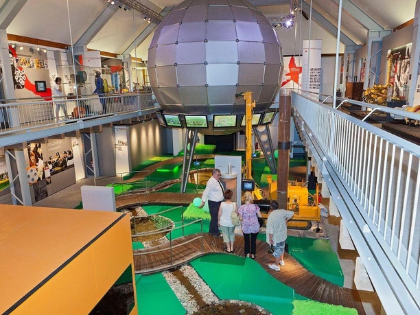 A museum with a large sphere, green floor and visitors on beams.