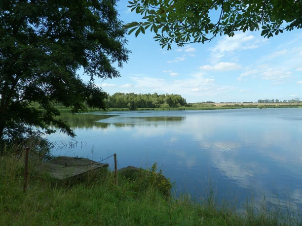 Klidná voda se stromy na břehu pod modrou oblohou, která se odráží ve vodě.