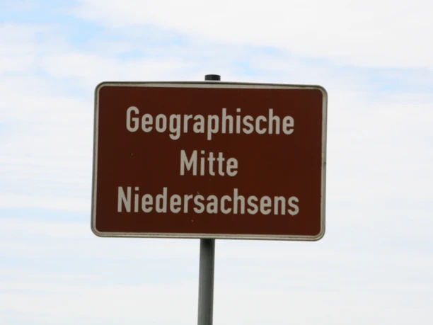 Ein Schild mit der Aufschrift "Geographische Mitte Niedersachsens" steht vor bewölktem Himmel.