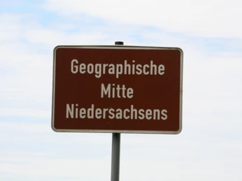 Geografischer Mittelpunkt Niedersachsen Ein Schild mit der Aufschrift "Geographische Mitte Niedersachsens" steht vor bewölktem Himmel.