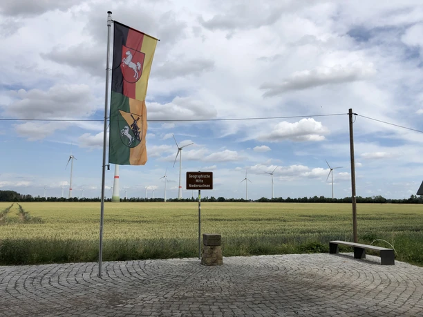 Geografischer Mittelpunkt Niedersachsens (16).jpg Vor einem windkraftbestückten Getreidefeld weht die Flagge Niedersachsens neben einem Wegweiser.
