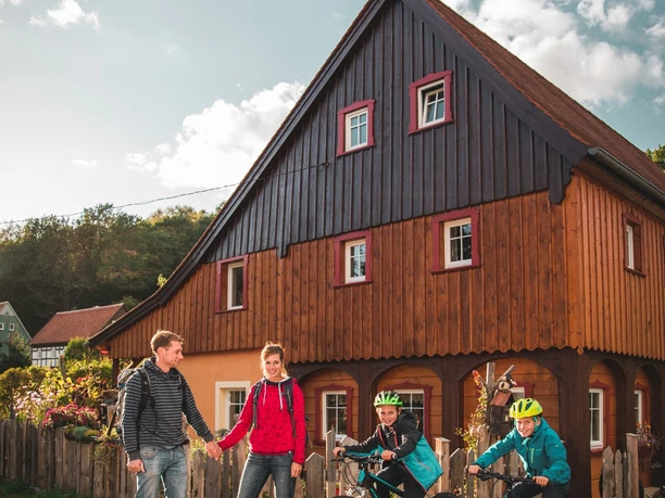 image Zwei Erwachsene und zwei Kinder mit Fahrrädern vor einem historischen Holzhaus bei Sonnenschein.