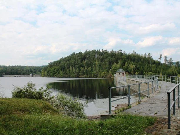 A path leads over a dam along a wooded lakeshore, surrounded by forest.