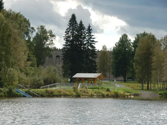 Vor der Staumauer der Talsperre Muldenberg Eine hölzerne Schutzhütte mit Blick auf eine bewaldete Staumauer und einen See.A wooden shelter with a view of a wooded dam wall and a lake.Dřevěný přístřešek s výhledem na zalesněnou hráz a jezero.Drewniana wiata z widokiem na zalesioną ścianę tamy i jezioro.Een houten schuilhut met uitzicht op een beboste damwand en een meer.Un rifugio in legno con vista su una parete di diga boscosa e su un lago.