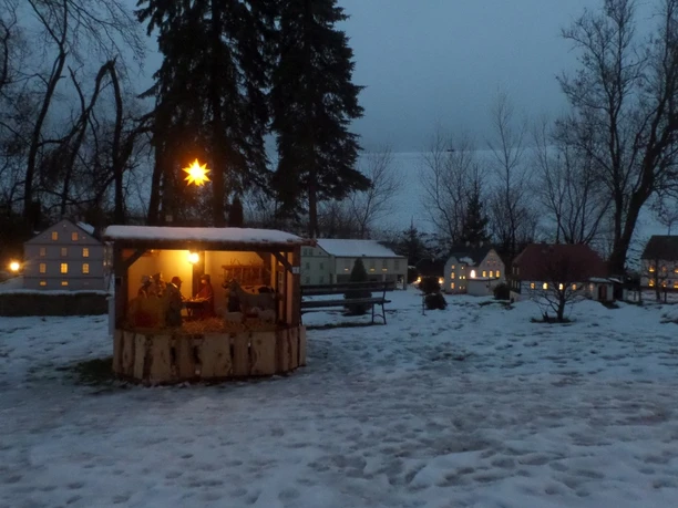 Mini-Weißbach Verschneites Miniaturdorf bei Dämmerung, beleuchtet mit Sternlichtern und Krippenszene.