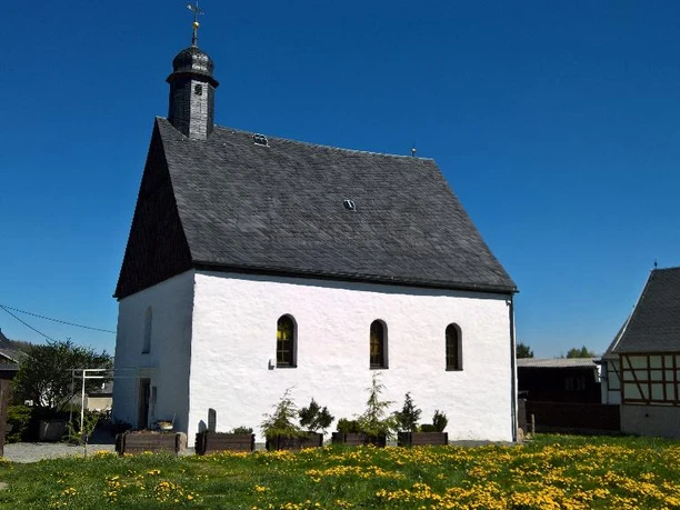 White chapel with slate roof surrounded by greenery, blue sky, half-timbered house next to it.