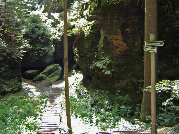 Dřevěná lávka vede stinným, zalesněným kamenitým potokem; směrovka ukazuje na Teufelsgrund.
