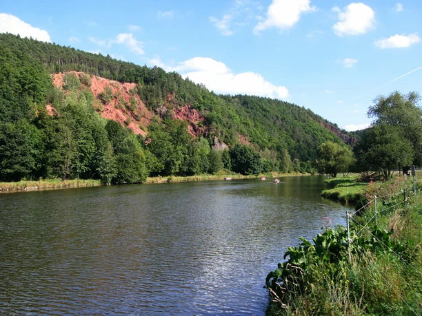 image Flussufer mit bewaldeten Hügeln und roten Felswänden, Boote gleiten über das Wasser.