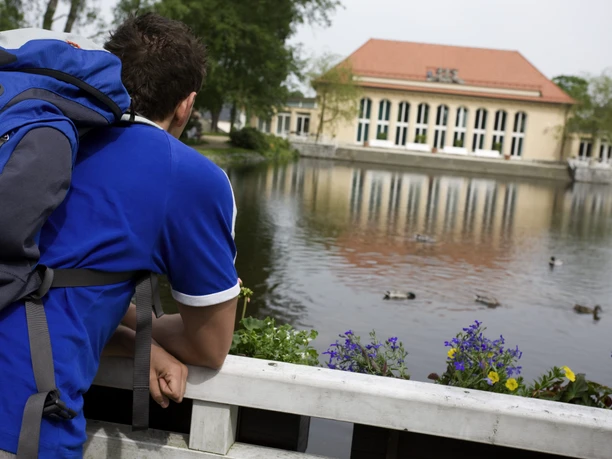 image Ein Mann mit blauem Rucksack betrachtet Enten auf einem Teich vor einem Gebäude im Hintergrund.
