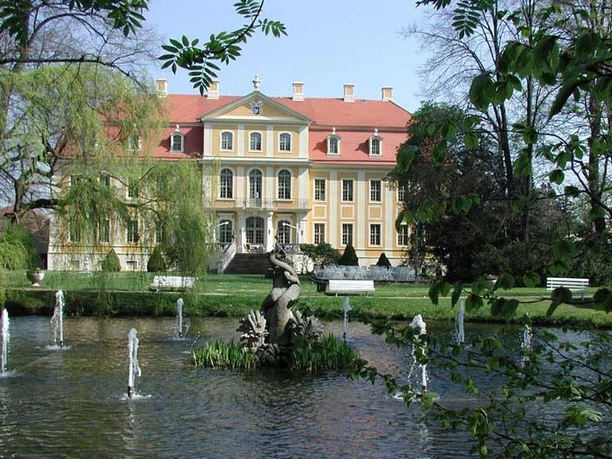 Historic castle with red roof and pond in the foreground, surrounded by trees.