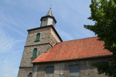 Alte Kirche mit rotem Ziegeldach und Turm mit Uhr unter einem blauen Himmel, umgeben von Bäumen.