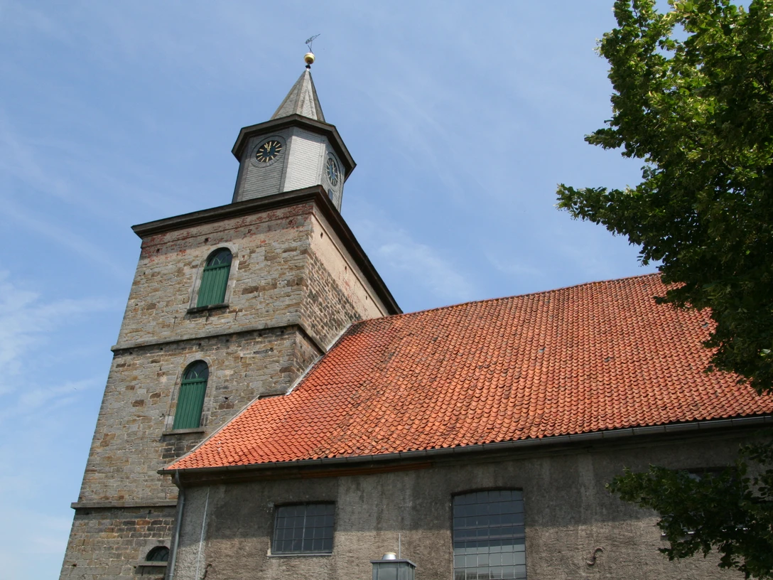 Alte Kirche mit rotem Ziegeldach und Turm mit Uhr unter einem blauen Himmel, umgeben von Bäumen.