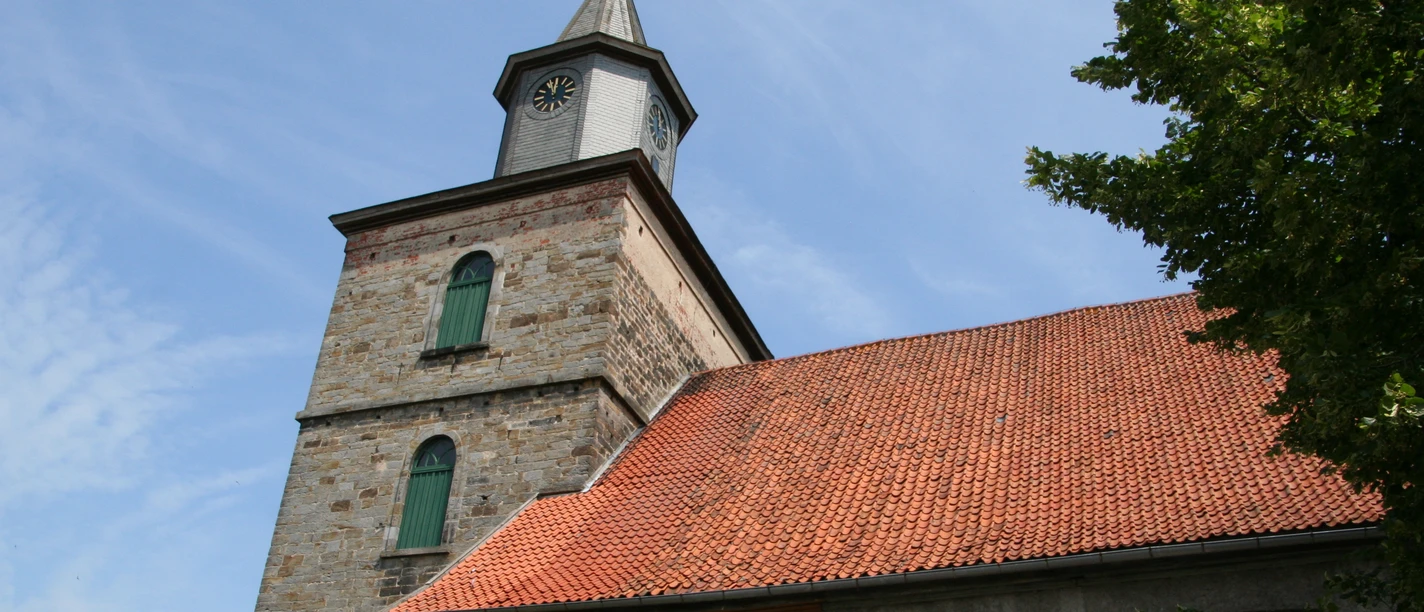 Alte Kirche mit rotem Ziegeldach und Turm mit Uhr unter einem blauen Himmel, umgeben von Bäumen.