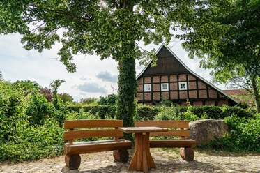 Nortrup im Artland Holzbank und Tisch im Schatten großer Bäume vor einem Fachwerkhaus an einem sonnigen Tag.