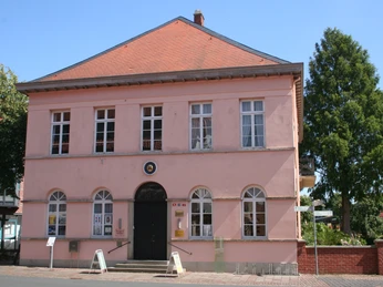 Quaet-Faslem-Haus Zweistöckiges rosa Gebäude mit Steildach, großen Fenstern und markanter Eingangstür, von Bäumen umgeben.Two-storey pink building with pitched roof, large windows and striking entrance door, surrounded by trees.Rosa bygning i to etager med skråt tag, store vinduer og markant indgangsdør, omgivet af træer.Roze gebouw van twee verdiepingen met schuin dak, grote ramen en opvallende ingangsdeur, omringd door bomen.