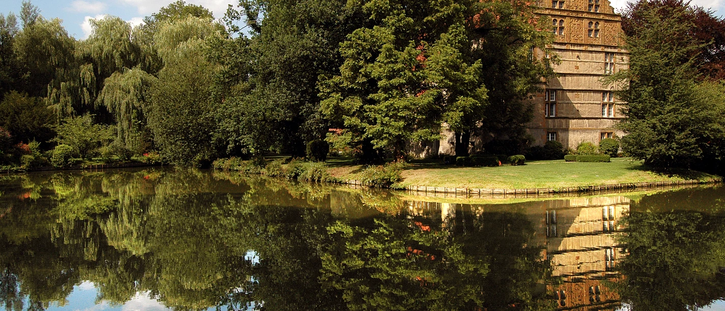 Wasserschloß Holtfeld Historisches Haus mit rotem Dach und Bäumen spiegelt sich idyllisch in einem ruhigen Teich wider.
